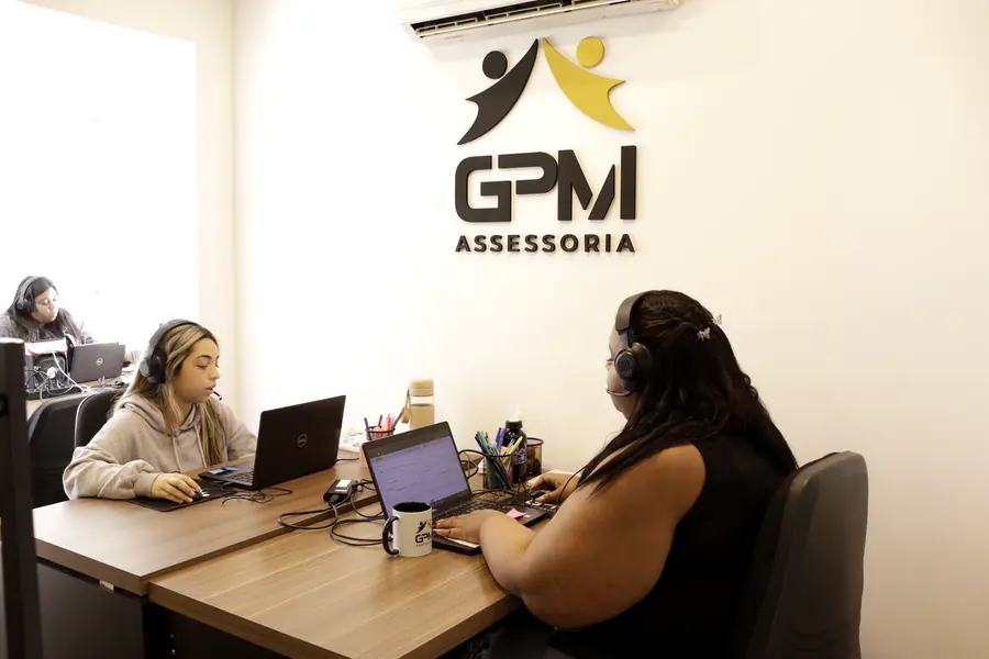Atendentes trabalhando em frente ao logo da GPM Assessoria na parede do escritório